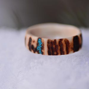 Könnte beinhalten: Ein handgefertigter Ring mit einer natürlichen, hellen Basis und dunkelbraunen, gestreiften Mustern. Ein kleiner Abschnitt aus leuchtend türkisfarbenem Inlay setzt einen Farbtupfer. Der Ring ist ein einzigartiges Schmuckstück.