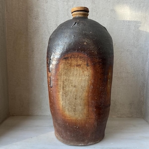 May include: A tall, antique stoneware jug with a dark brown, glazed top and a lighter brown, unglazed body. The jug has a rounded shoulder and a small, wooden stopper. The overall appearance is rustic and aged, with visible imperfections.