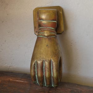 Puede incluir: Un llamador de puerta de latón con forma de puño cerrado. El metal tiene un acabado envejecido y desgastado con oxidación verde visible en los pliegues de los dedos y la muñeca. Incluye una placa de montaje rectangular en la parte superior.