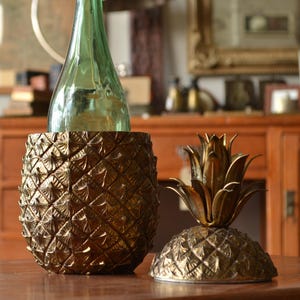 Puede incluir: Un soporte para botellas de vino en forma de piña de color dorado con una botella de vidrio verde en su interior. Un tapón de botella a juego en forma de piña está al lado. Los objetos están sobre una mesa de madera.