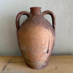 Op de afbeelding: Een rustieke terracotta amfora met twee handvatten en een verweerde uitstraling. Het vat heeft een bolvormige body en een smalle hals, met twee gebogen handvatten. De kleur is een mix van roodbruin en bruin, met tekenen van slijtage.