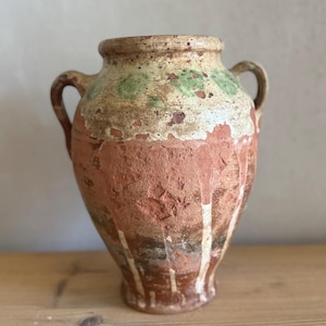 Puede incluir: Un jarrón de terracota antiguo con dos asas. El jarrón presenta un aspecto envejecido y desgastado, con capas de pintura roja, crema y verde. El jarrón probablemente se utiliza con fines decorativos. Altura aproximada 30 cm.
