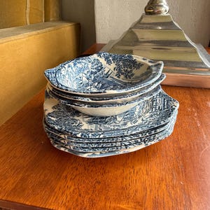 Può includere: Una pila di piatti e ciotole in ceramica blu e bianca con un design paesaggistico. I piatti sono ovali e impilati su una superficie di legno. Il design presenta una scena paesaggistica. I piatti e le ciotole sono in stile classico.