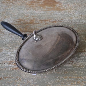 Peut inclure: Plat de service ovale argenté avec une poignée noire. Le plat a une bordure décorative en forme de corde et une petite poignée ornée sur le couvercle. Le fond est une surface en bois clair vieilli.