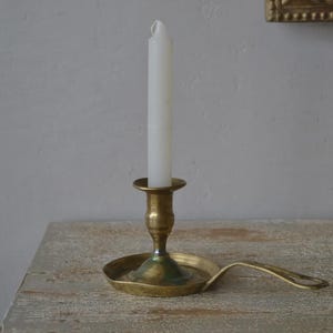 May include: A brass candlestick with a white candle. The candlestick has a round base with a handle and a central holder for the candle. The candle is white and has a slightly melted top. The candlestick is on a wooden surface.