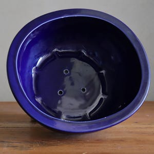 Könnte beinhalten: Ein tiefblauer Keramik-Pflanztopf mit glänzender Oberfläche. Der runde Topf hat eine weite Öffnung und eine leicht konische Form. Drei Abzugslöcher sind am Boden sichtbar. Der Topf steht auf einer Holzoberfläche.