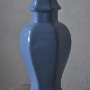 Może przedstawiać: Jasnoniebieski ceramiczny wazon o unikalnym, geometrycznym kształcie. Wazon ma rozszerzoną podstawę, smukły korpus i zaokrągloną g&oacute;rę z małą, płaską pokrywką. Błyszczące wykończenie odbija światło. Wazon ma około 30 cm wysokości.