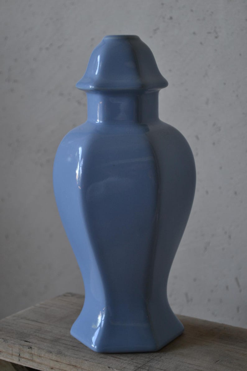Może przedstawiać: Jasnoniebieski ceramiczny wazon o unikalnym, geometrycznym kształcie. Wazon ma rozszerzoną podstawę, smukły korpus i zaokrągloną g&oacute;rę z małą, płaską pokrywką. Błyszczące wykończenie odbija światło. Wazon ma około 30 cm wysokości.