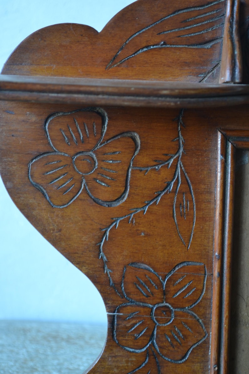 May include: Close-up of a wooden shelf with carved floral designs. The shelf is made of a warm-toned wood and features two carved flowers and a stem with leaves. The shelf has a scalloped edge.