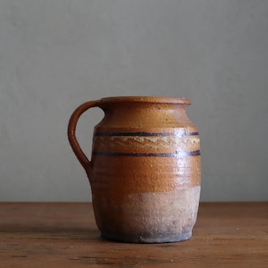 Op de afbeelding: Een rustieke keramische kruik met een handvat. De kruik is voornamelijk bruin met lichtere beige en crèmetinten. Hij heeft twee donkerblauwe horizontale strepen en een decoratieve band nabij de bovenkant. Hij staat op een houten oppervlak.