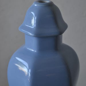 Może przedstawiać: Jasnoniebieski ceramiczny wazon z unikalnym, rozszerzanym topem. Wazon ma gładkie, błyszczące wykończenie i zaokrąglony korpus o lekko kanciastym kształcie. Wazon ma około 25 cm wysokości.