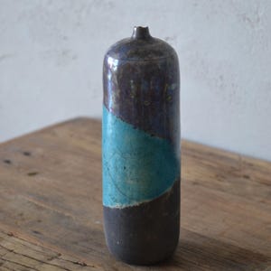Op de afbeelding: Een cilindrische keramische vaas met een smalle hals en een afgeronde bovenkant. Het oppervlak heeft een donker, gevlekt bruin glazuur op de boven- en onderkant, gecontrasteerd door een levendige, gladde turquoise band in het midden met fijne lijntjes.
