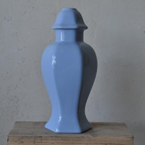 Może przedstawiać: Jasnoniebieski ceramiczny wazon z pokrywką. Wazon ma błyszczące wykończenie i geometryczny, fasetowany design. Jest prezentowany na drewnianej powierzchni na neutralnym tle.
