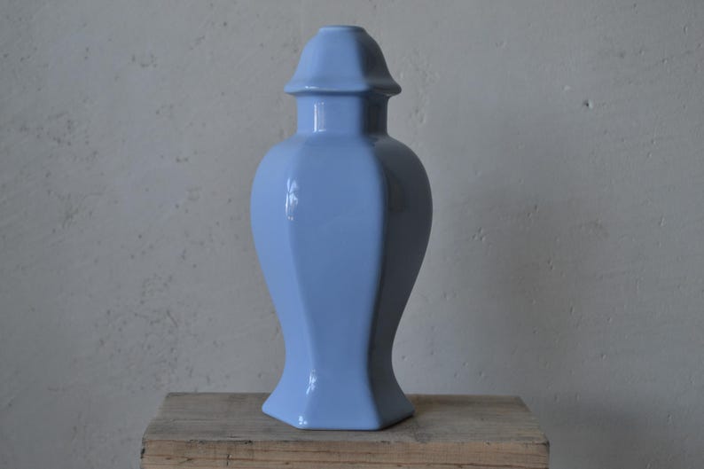 Może przedstawiać: Jasnoniebieski ceramiczny wazon z pokrywką. Wazon ma błyszczące wykończenie i geometryczny, fasetowany design. Jest prezentowany na drewnianej powierzchni na neutralnym tle.