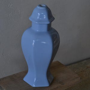 Może przedstawiać: Jasnoniebieski ceramiczny wazon z pokrywką, ustawiony na rustykalnym drewnianym stołku. Wazon ma błyszczące wykończenie i unikalny, geometryczny kształt. Tło to neutralna szara ściana.