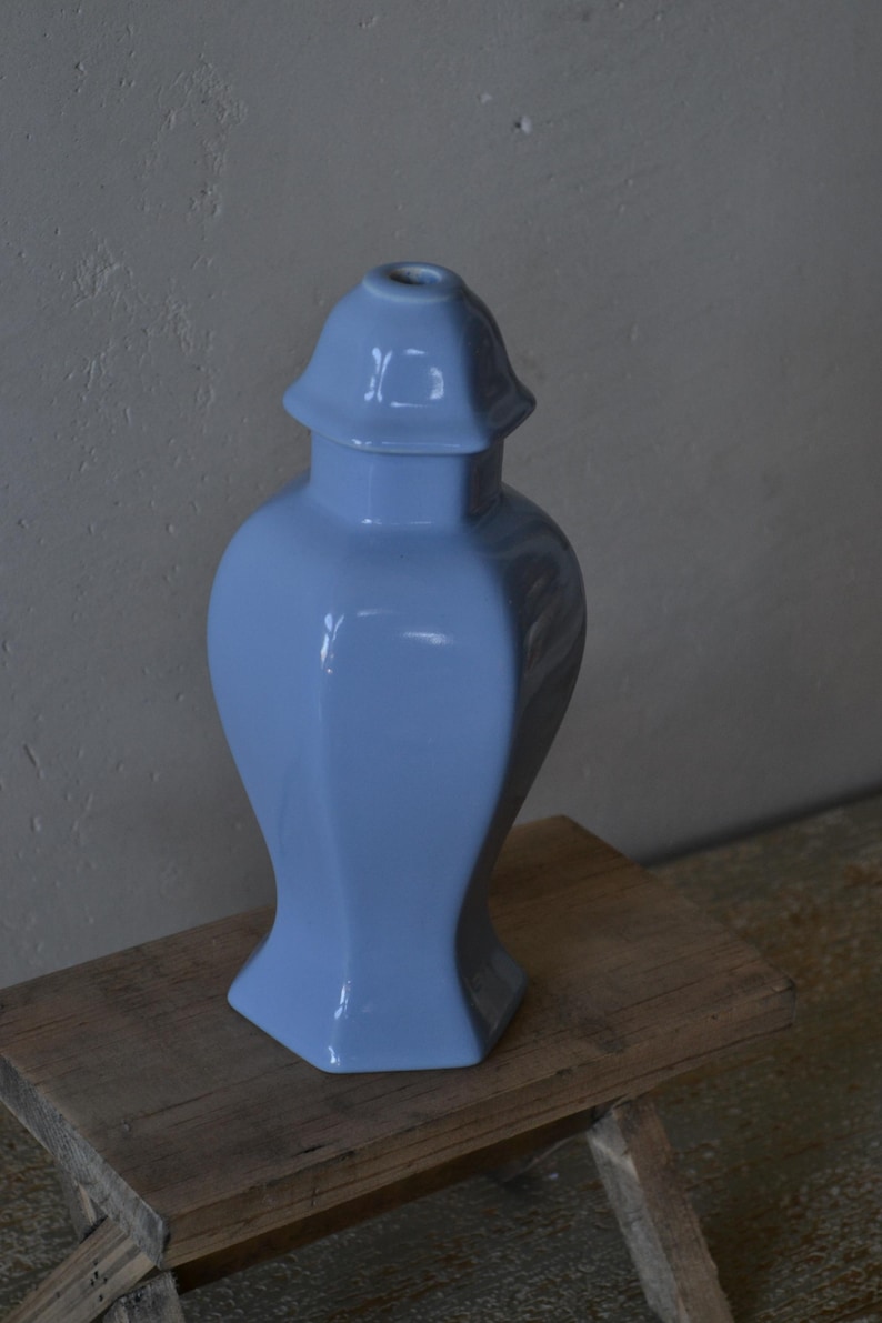 Może przedstawiać: Jasnoniebieski ceramiczny wazon z pokrywką, ustawiony na rustykalnym drewnianym stołku. Wazon ma błyszczące wykończenie i unikalny, geometryczny kształt. Tło to neutralna szara ściana.