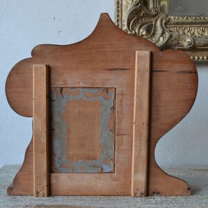 May include: Back view of a vintage wooden picture frame. The frame is made of light brown wood with a decorative top and base. The back has a rectangular opening with remnants of old paper and a support structure.