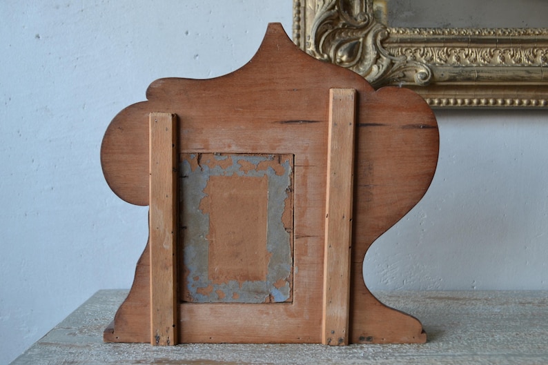 May include: Back view of a vintage wooden picture frame. The frame is made of light brown wood with a decorative top and base. The back has a rectangular opening with remnants of old paper and a support structure.