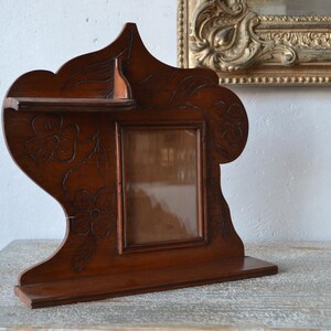 May include: A dark brown wooden picture frame with a shelf and floral carvings. The frame has a rectangular opening for a photo and is set on a base. A decorative piece for displaying photos.