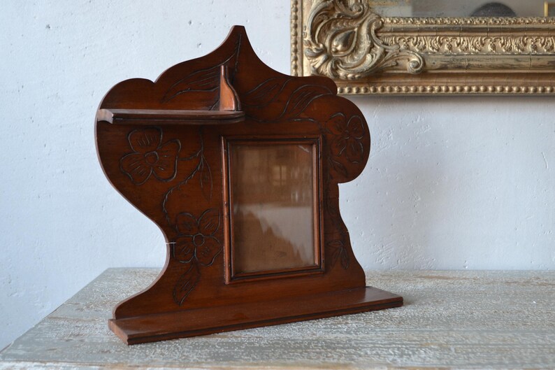 May include: A dark brown wooden picture frame with a shelf and floral carvings. The frame has a rectangular opening for a photo and is set on a base. A decorative piece for displaying photos.