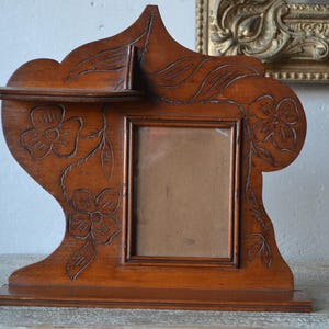May include: A wooden picture frame with a shelf and floral carvings. The frame is a warm brown color and features a rectangular opening for a photo. The floral carvings are on the sides and top of the frame.