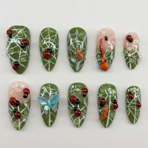 May include: A set of ten artificial nails with a variety of designs. The nails feature a green base with white web-like patterns and red ladybugs. Some nails have orange insects, a blue butterfly, and clear droplets.