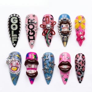 May include: A collection of ten long, pointed artificial nails with various designs. The nails feature embellishments like rhinestones, text, and 3D elements, including lips, flowers, and cartoon faces. Colors include black, pink, blue, and leopard print.