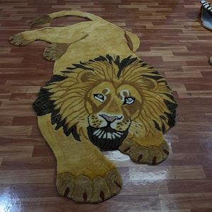 May include: A custom-shaped area rug featuring a detailed, tufted lion design in shades of golden yellow, tan, and dark brown. The lion is depicted in a reclining position with its head facing forward, showcasing a full mane and textured paws.