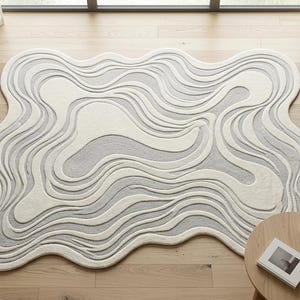 Op de afbeelding: Een bovenaanzicht van een modern, abstract vloerkleed met een golvend ontwerp in grijstinten en crème. Het kleed, ongeveer 213 x 152 cm, ligt op een lichte houten vloer. Een bank, salontafel en een plant in een pot zijn zichtbaar.