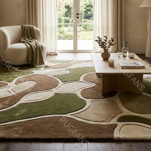 May include: A large area rug with an abstract design in shades of green, brown, and beige. The rug is placed in a living room setting with a sofa, armchair, and coffee table. The rug's pattern is organic and modern.