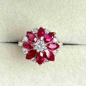 Könnte beinhalten: Ein silberner Ring mit floralem Design. Das Zentrum des Rings zeigt eine Ansammlung kleiner, runder weißer Steine, umgeben von leuchtend roten, blütenblattförmigen Steinen. Der Ring wird in einer Schmuckschatulle präsentiert.