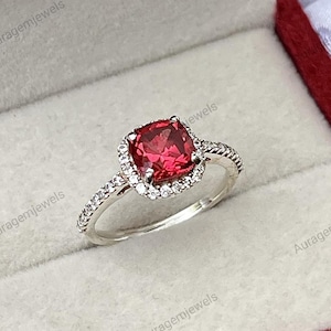 May include: A silver ring featuring a square, red gemstone encircled by a halo of smaller, clear stones. The ring is presented in a red velvet box, with the brand name "Auragemjewels" visible.