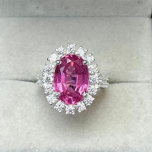 Puede incluir: Un anillo de piedra preciosa rosa con un halo de pequeñas piedras transparentes. El anillo está engastado en una banda plateada y se muestra en una caja de joyería marrón con un interior gris. La piedra rosa de forma ovalada es el punto focal.