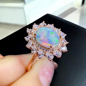 May include: Rose gold ring featuring a large, oval opal gemstone surrounded by a halo of smaller, clear gemstones. The opal displays iridescent colors, and the ring's band is slender.