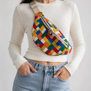 May include: A colorful fanny pack with a pattern resembling toy building blocks. The bag features a zipper closure and a beige strap. The design incorporates red, yellow, blue, green, and orange blocks. The bag is worn across the body.