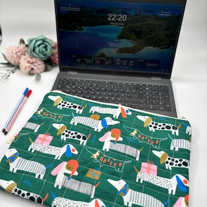 May include: Green laptop sleeve featuring a repeating pattern of dachshund dogs in various colors and designs. The dogs have hats, gifts, and other accessories. The sleeve is on a white surface with a laptop and pens in the background.