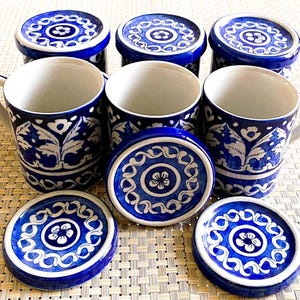 May include: A set of blue and white ceramic mugs and matching coasters. The mugs have a floral design and handles. The coasters and lids have a circular design with a flower in the center. The set includes six mugs, six lids, and three coasters.