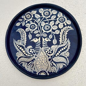 Op de afbeelding: Een ronde, marineblauwe decoratieve schaal met een wit bloemen- en vogelontwerp. De schaal heeft een gedetailleerd patroon van bloemen, bladeren en twee gestileerde vogels. De rand van de schaal is effen marineblauw.