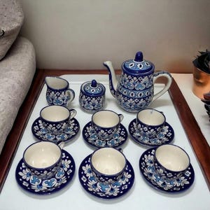 May include: A ceramic tea set featuring a teapot, a lidded sugar bowl, a creamer, and six cups with matching saucers. Each piece is decorated with a blue and white floral pattern on a cream-coloured background, showcasing intricate hand-painted details.