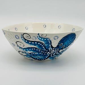 May include: A white ceramic bowl with a textured exterior and a blue octopus design. The octopus is painted with intricate details, including white dots and swirling tentacles. Blue bubbles are scattered around the rim.