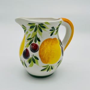 Può includere: Brocca in ceramica bianca con manico e beccuccio gialli. La brocca è decorata con limoni gialli, ciliegie rosso scuro e foglie verdi. La base della brocca è verde.