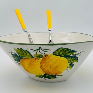 May include: A white ceramic serving bowl with a green rim and a textured exterior. The bowl is decorated with yellow lemons and green leaves. Two yellow-handled serving utensils are inside the bowl.