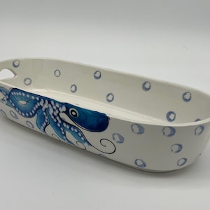 Può includere: Piatto da portata ovale in ceramica bianca con design a polpo blu. Il piatto presenta un polpo dipinto a mano su un lato e pois blu attorno al bordo. Ha due piccole maniglie ed è adatto per servire snack o piccoli piatti.