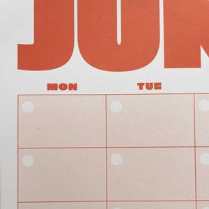 May include: Close-up of a calendar page with the month of June in large orange letters. The days of the week, "MON" and "TUE", are in smaller orange letters. The calendar grid has light pink boxes with white circles.
