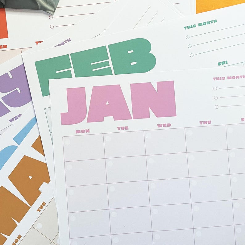 May include: A stack of colorful monthly planner sheets. The sheets feature large, bold text for the months of the year, including "JAN" in pink and "FEB" in green. Each sheet has a grid for daily planning and the words "THIS MONTH" at the top.