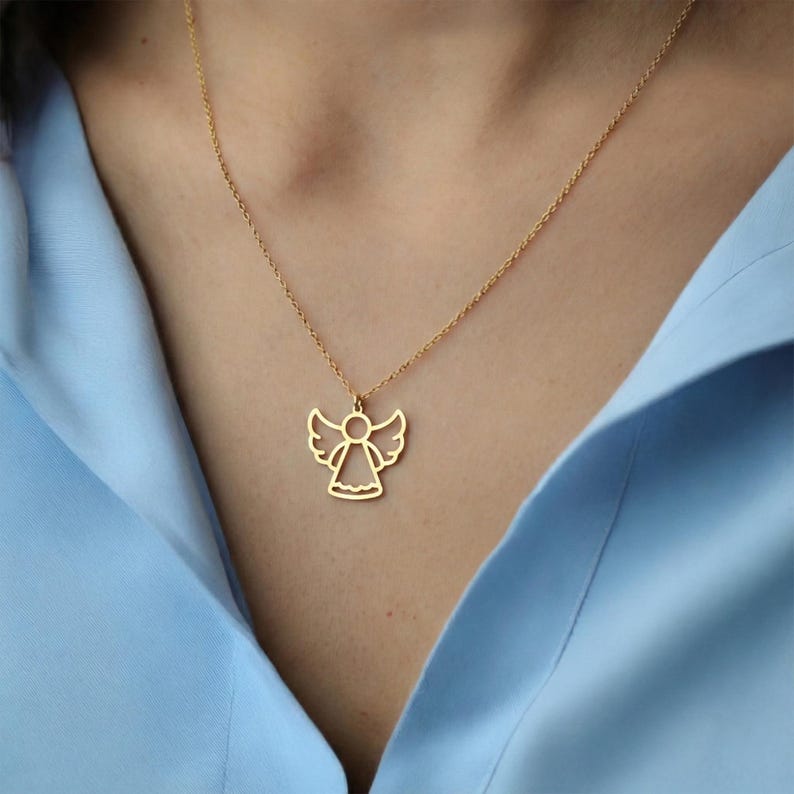 May include: A gold-colored angel pendant necklace. The pendant features a stylized angel with outstretched wings and a flowing gown. The necklace is worn against a light blue shirt.
