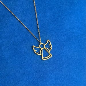 May include: A gold-colored angel pendant necklace. The pendant features a stylized angel outline with outstretched wings and a flowing skirt. The necklace chain is also gold and hangs against a blue background.