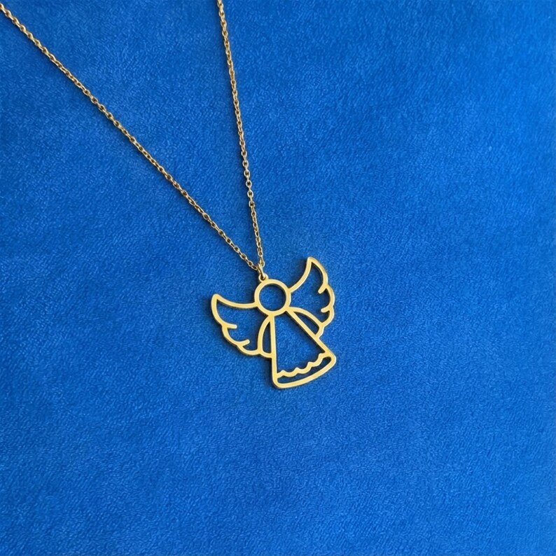 May include: A gold-colored angel pendant necklace. The pendant features a stylized angel outline with outstretched wings and a flowing skirt. The necklace chain is also gold and hangs against a blue background.