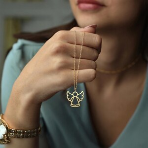 May include: A gold-colored necklace featuring an angel pendant. The pendant has a cutout design with wings and a halo. The necklace is held up by a person, showcasing the delicate chain and the pendant's details. The background is blurred.