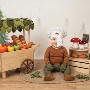 May include: A wooden toy market cart filled with colorful toy fruits and bread. A baby wearing a chef's hat with a mushroom design sits in front of the cart. Decorative mushroom toys and a chalkboard are also present.